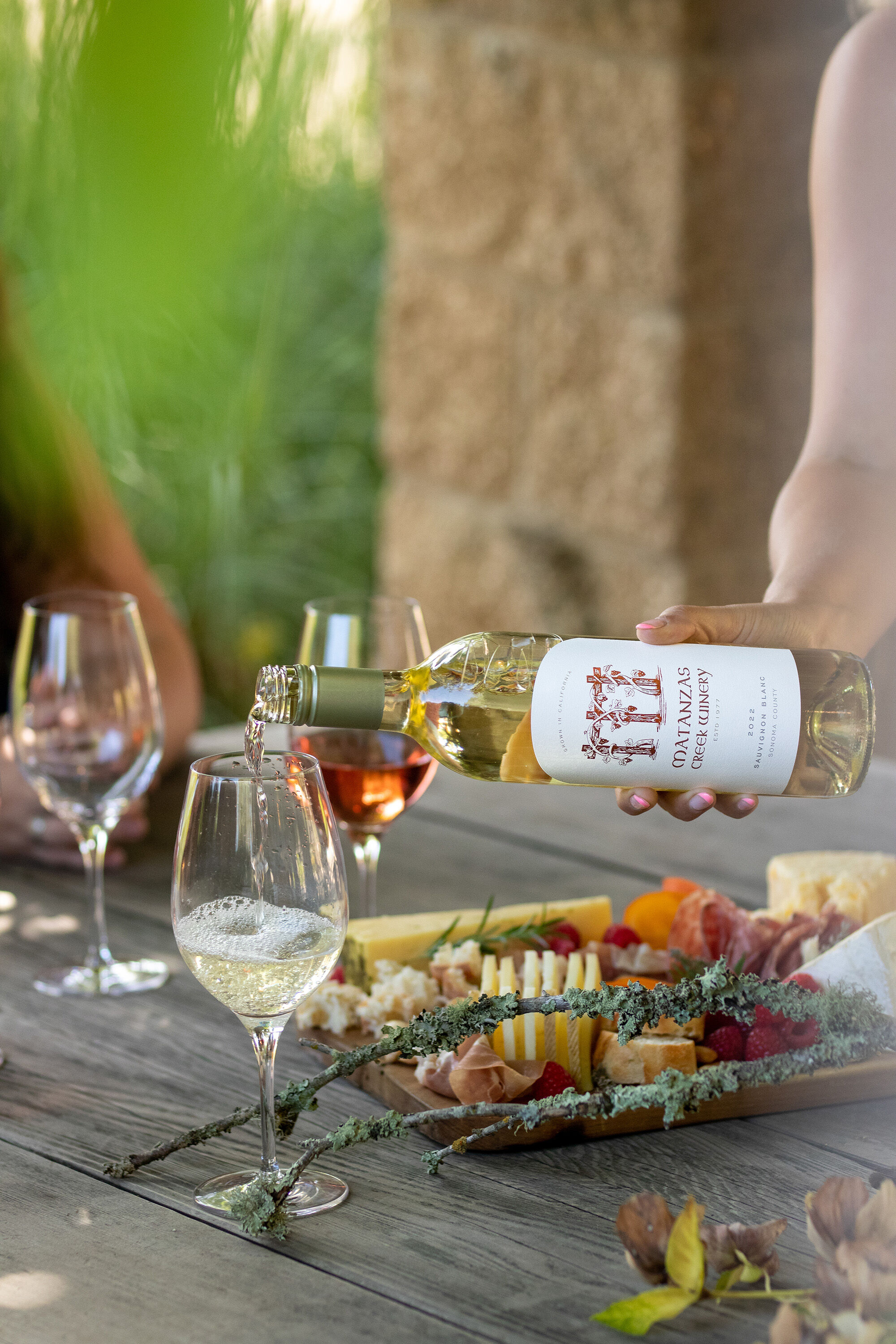 A close up of a white wine bottle pouring wine into a glass, with a cheese and charcuterie board.