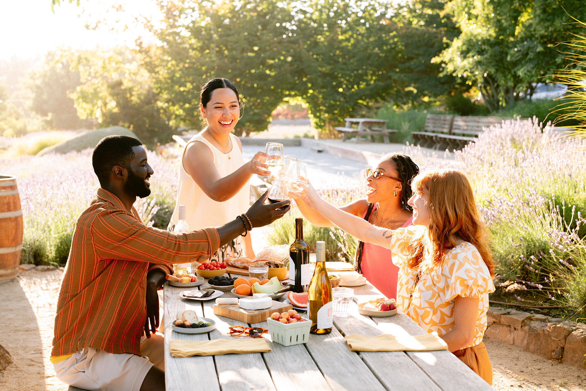 A group of four people around a picnic table with bottles of wine and food, who are smiling and clinking glasses.