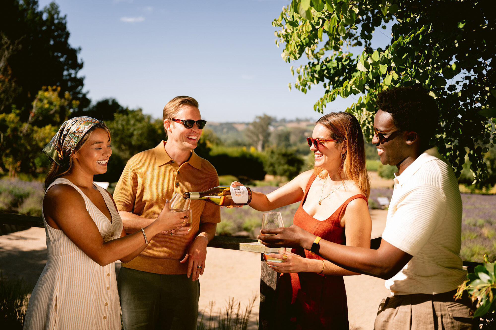 A group of two women and two men laughing and pouring a bottle of wine.