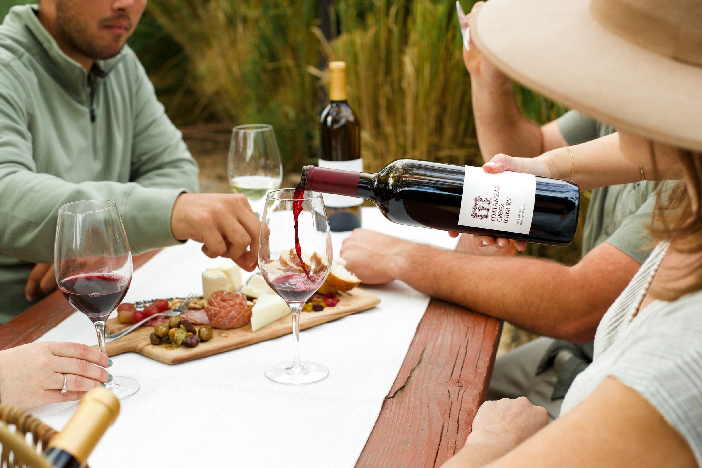 A group of three people at a picnic table eating a charcuterie board with wine glasses.