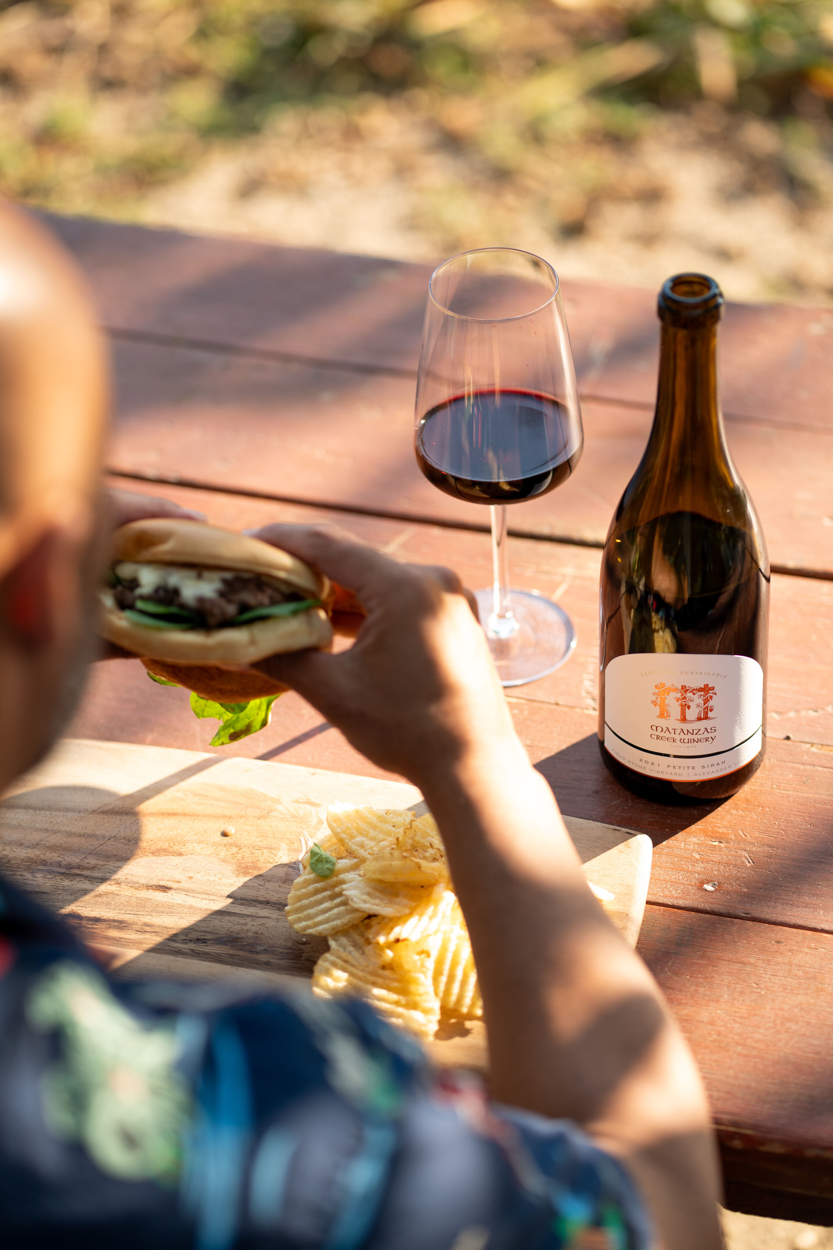 Outdoor image at a picnic bench of a person eating a burger with a bottle and glass of Matanzas Creek