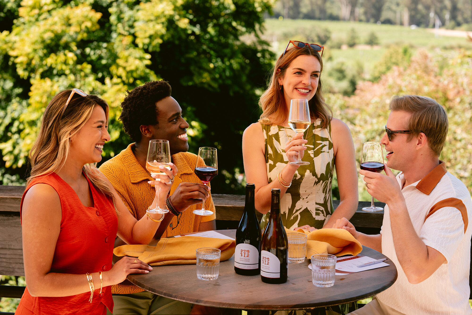 A group of two men and two women around a round wood table, smiling with glasses and bottles of wine.